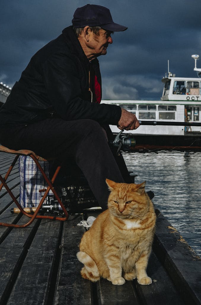 Elderly man fishing with a cat by the waterfront against a cloudy sky.