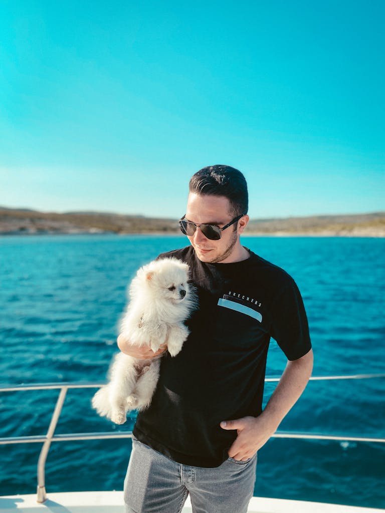 Man with sunglasses holds a dog on a yacht under a bright blue sky in Alaçatı, Türkiye.