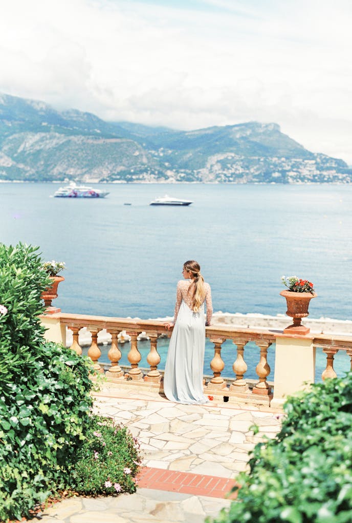 Woman in flowing dress gazes at scenic ocean view from elegant terrace with mountain backdrop.