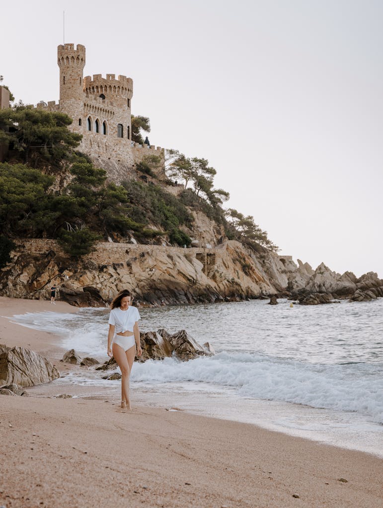 A woman walks along a sandy beach with a castle on the hill, surrounded by lush greenery and the sea.