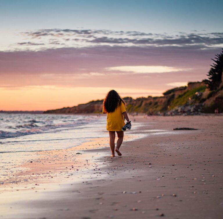 A woman walks barefoot along Pénestin Beach in a yellow dress, capturing the vibrant sunset over the sea.